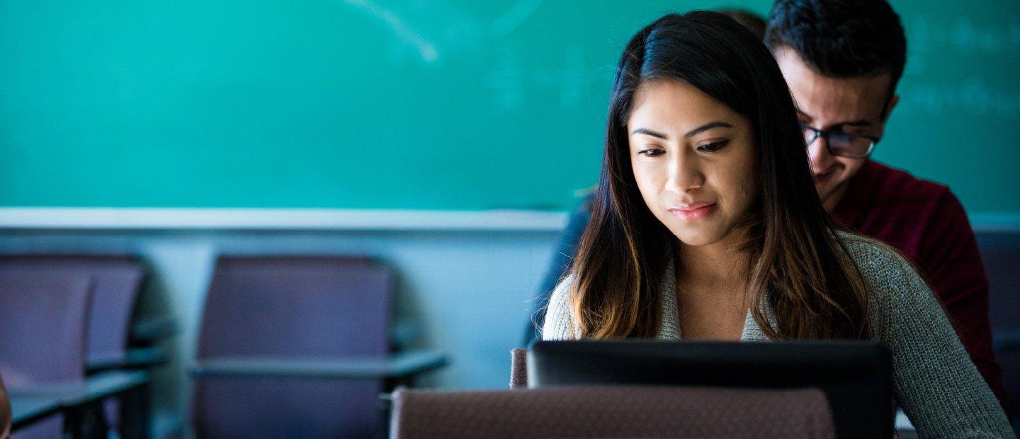 two students seated, looking at laptops, with a green chalkboard in the background